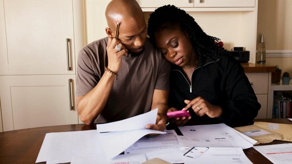 A couple sits at a table managing domestic finances, evaluating documents and using a smartphone.