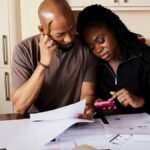 A couple sits at a table managing domestic finances, evaluating documents and using a smartphone.
