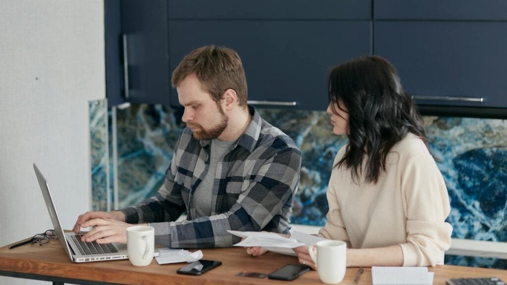A couple reviews bills and finances at a desk with a laptop and papers. Indoor setting, focused on budgeting.