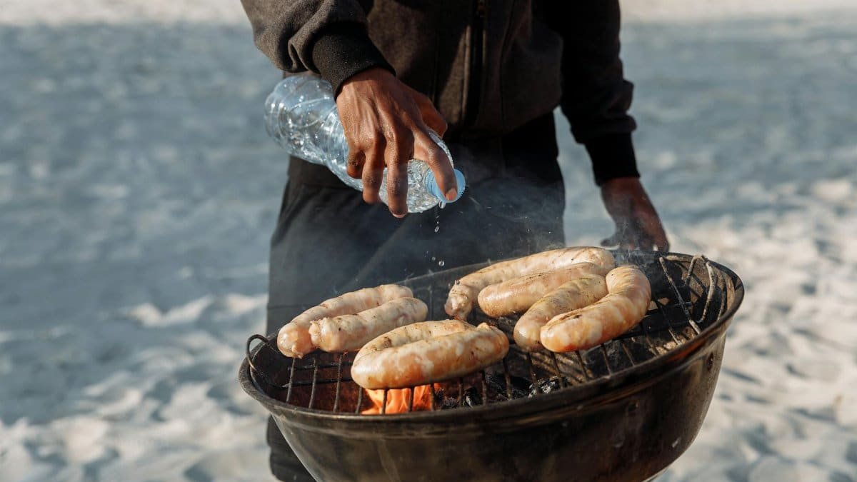 Person grilling sausages on a beach with a portable barbecue and bottled water.