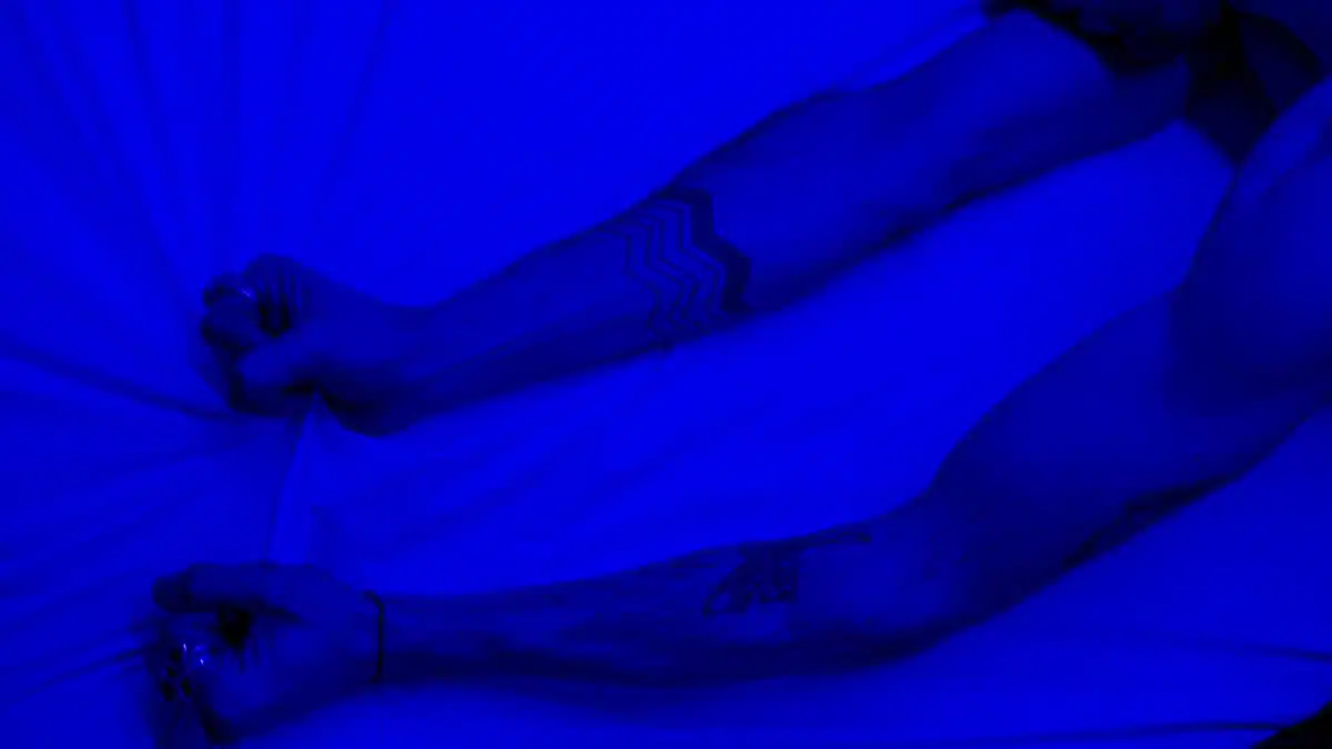 Close-up of tattooed arms grasping a white bedsheet under blue lighting, depicting anxiety and tension.