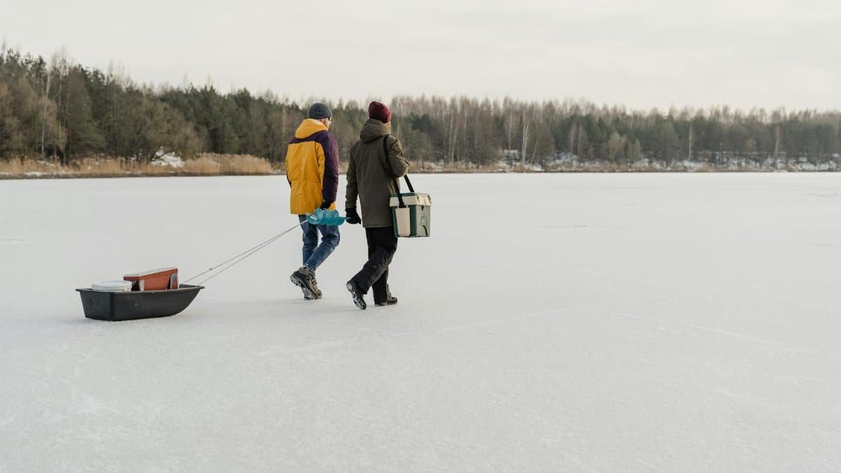 Two friends walk on a frozen lake, enjoying winter in outdoor attire, pulling a sled with gear.