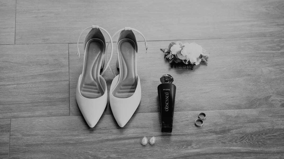 Black and white flat lay of bridal shoes, perfume, rings, and floral corsage on wooden floor.