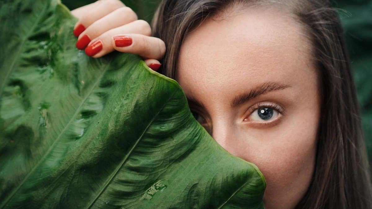 Close-up shot of a woman with red nails, partially hidden behind a large green leaf.