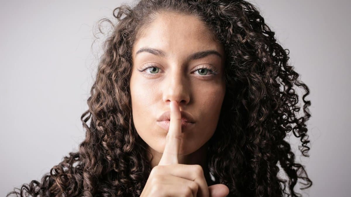 Close-up portrait of a woman with curly hair, signaling quiet with a finger on lips.