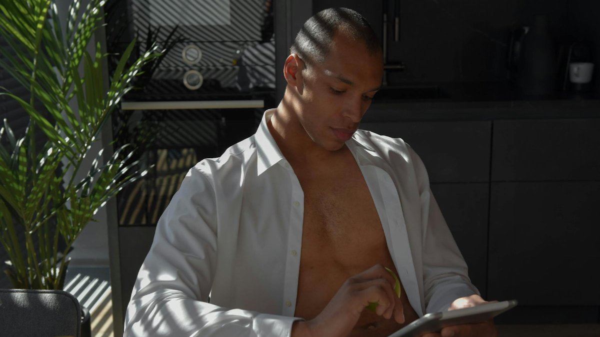 Young man in an open shirt using a tablet in a bright, sunlit kitchen setting.