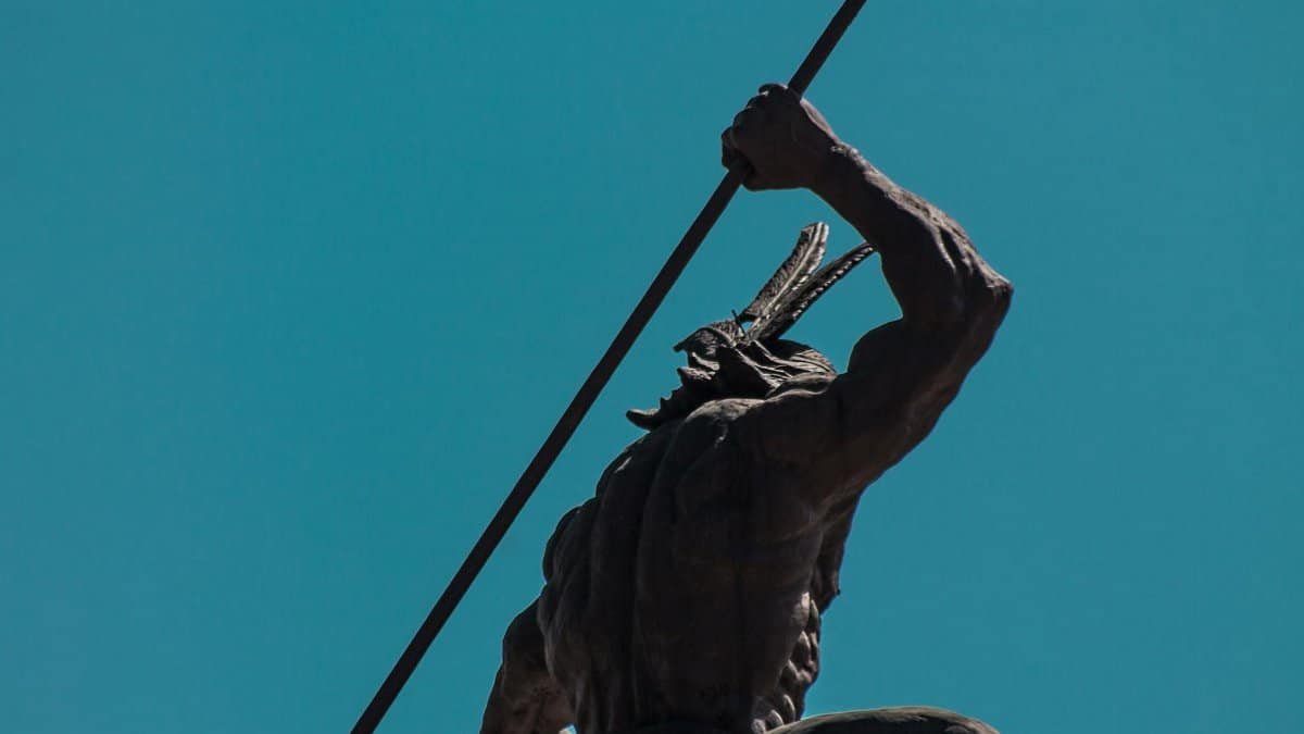 Low angle shot of an indigenous warrior statue with spear under a clear blue sky in a dynamic pose.