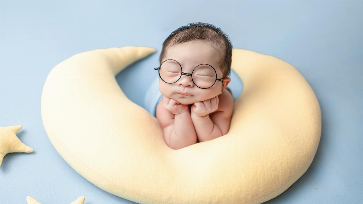 Charming portrait of a newborn baby with glasses cradled in a moon-shaped pillow.