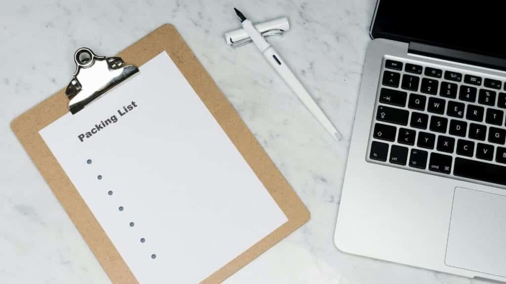 Flat lay of a packing list clipboard with a laptop and pen on a marble desk.