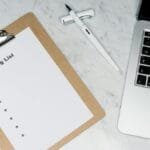 Flat lay of a packing list clipboard with a laptop and pen on a marble desk.