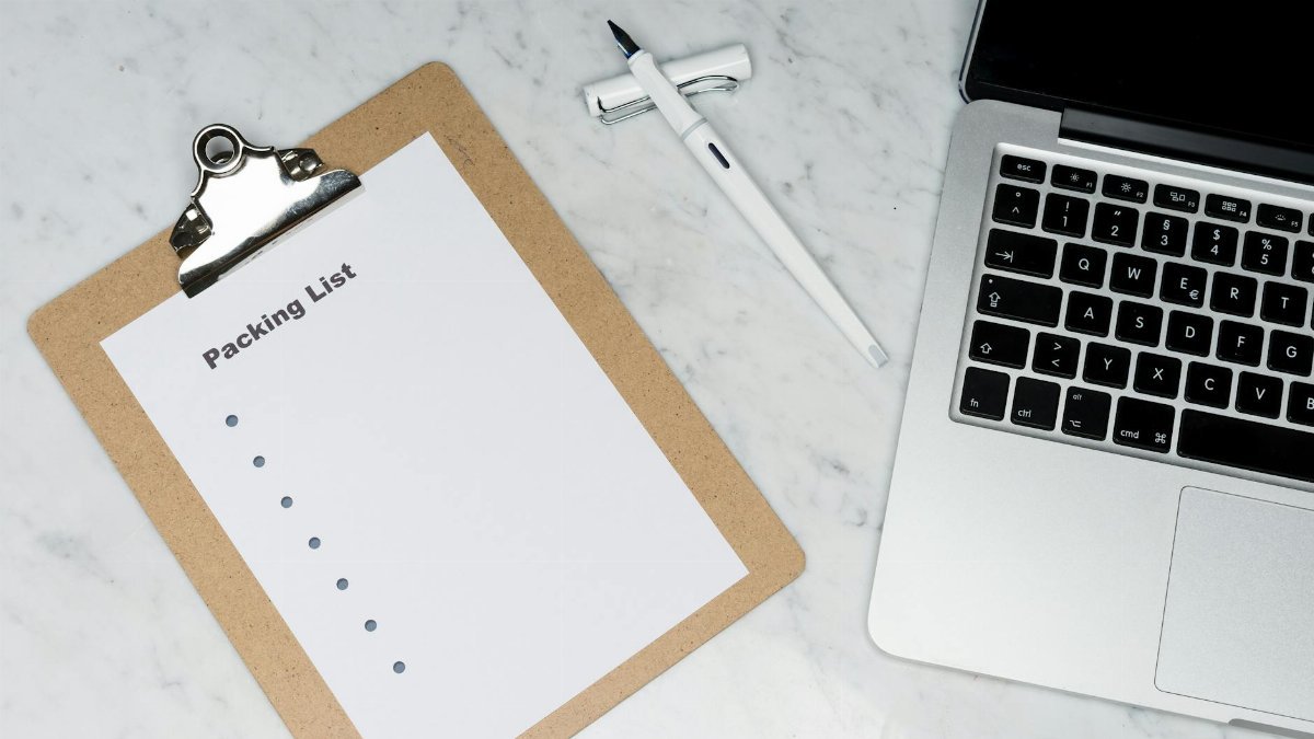Flat lay of a packing list clipboard with a laptop and pen on a marble desk.
