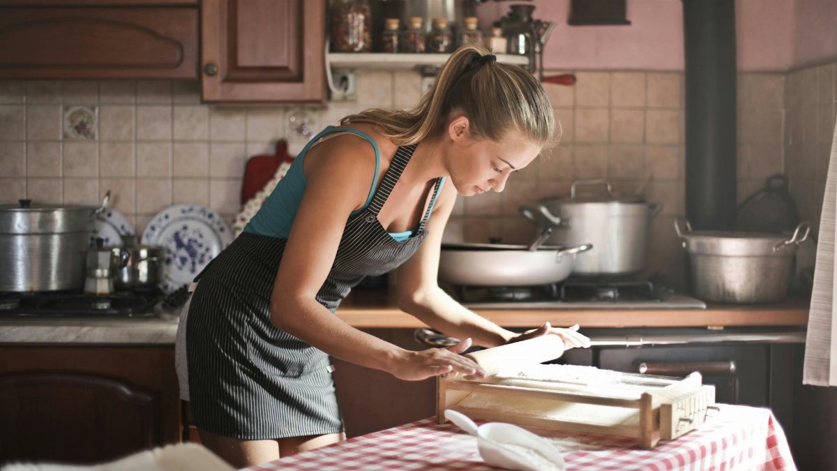 Side view of housewife wearing apron standing at table in cozy kitchen and preparing dough for baking while using rolling pin