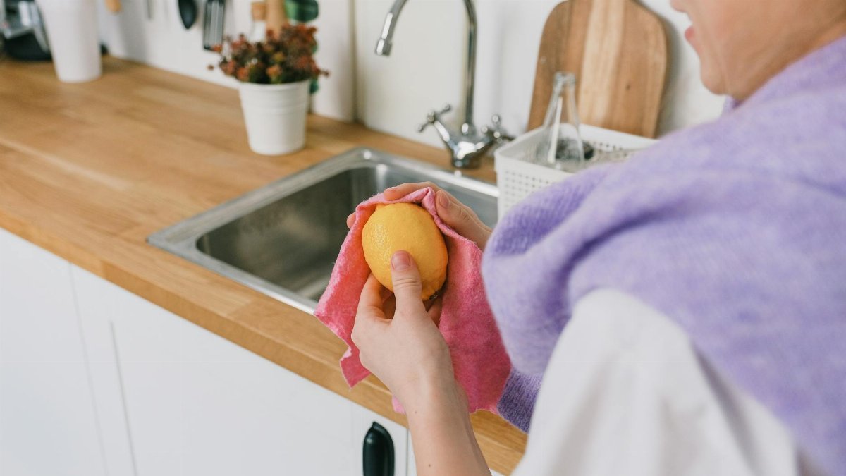 Crop unrecognizable person in casual clothes standing and wiping ripe fresh lemon with cloth in light kitchen in daytime