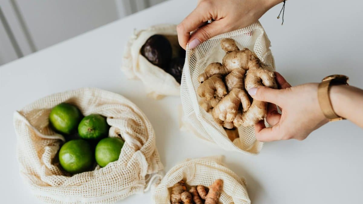 Hands sorting ginger, limes, avocados, and turmeric in reusable net bags on a white surface.