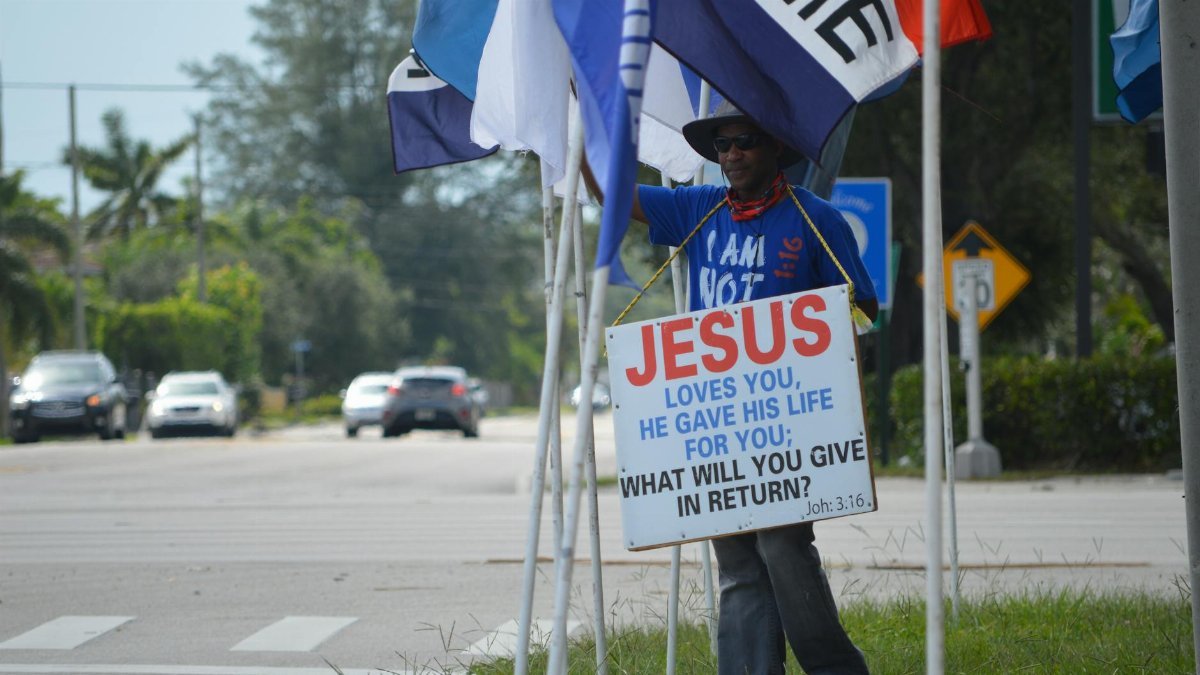 A man holding a religious sign with flags at a West Palm Beach intersection.