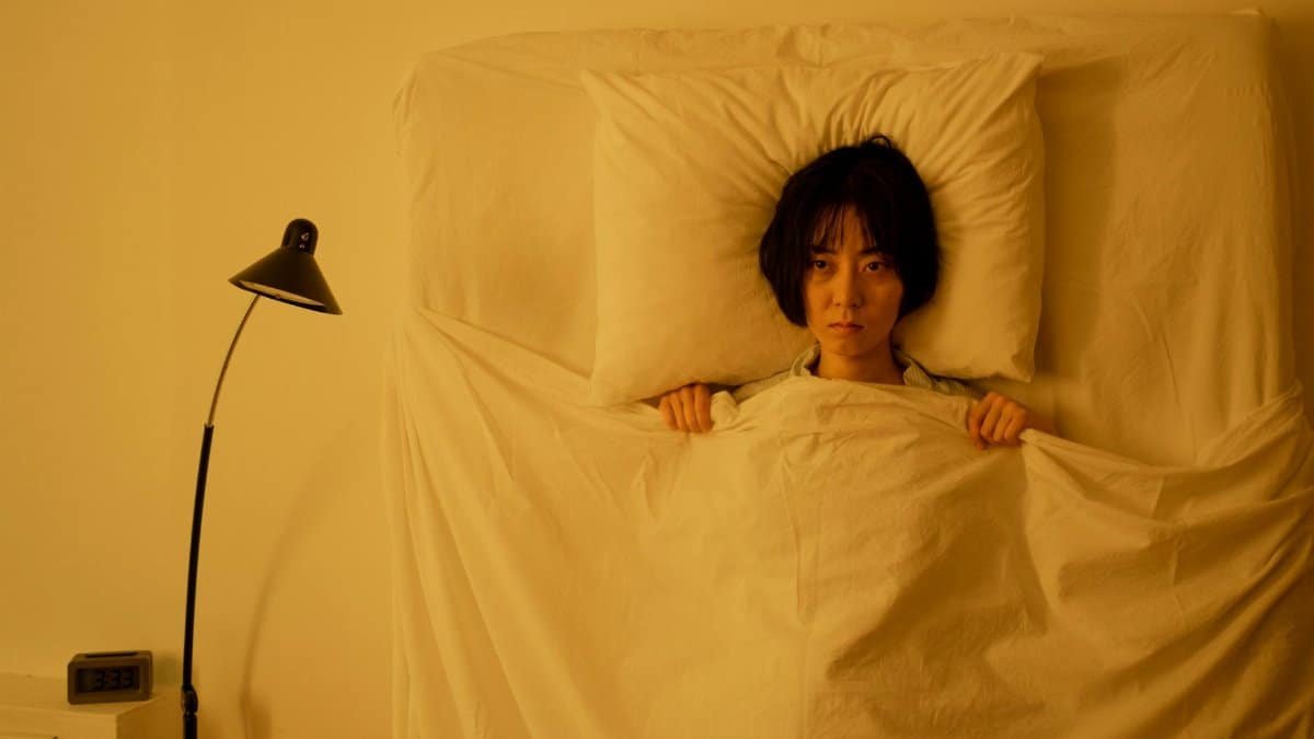 Woman lying under a blanket with a moody expression in a dimly-lit bedroom.