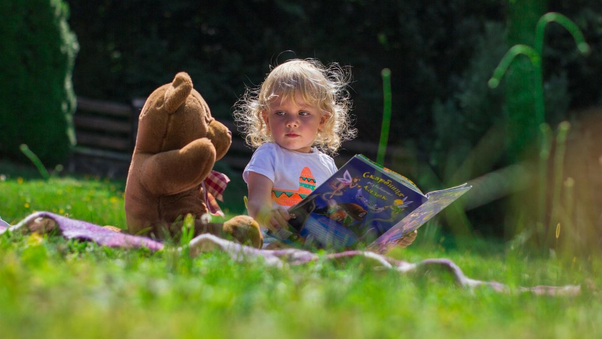 Young child reading a book outdoors with a teddy bear, enjoying a sunny day on the grass.