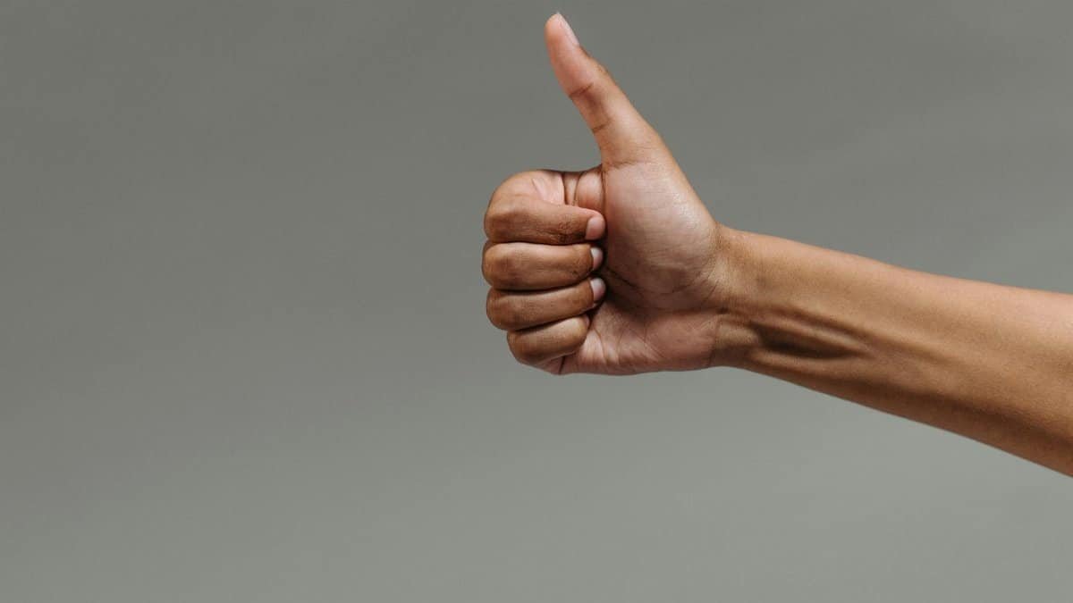 A close-up image of a hand making a thumbs up gesture against a neutral background.