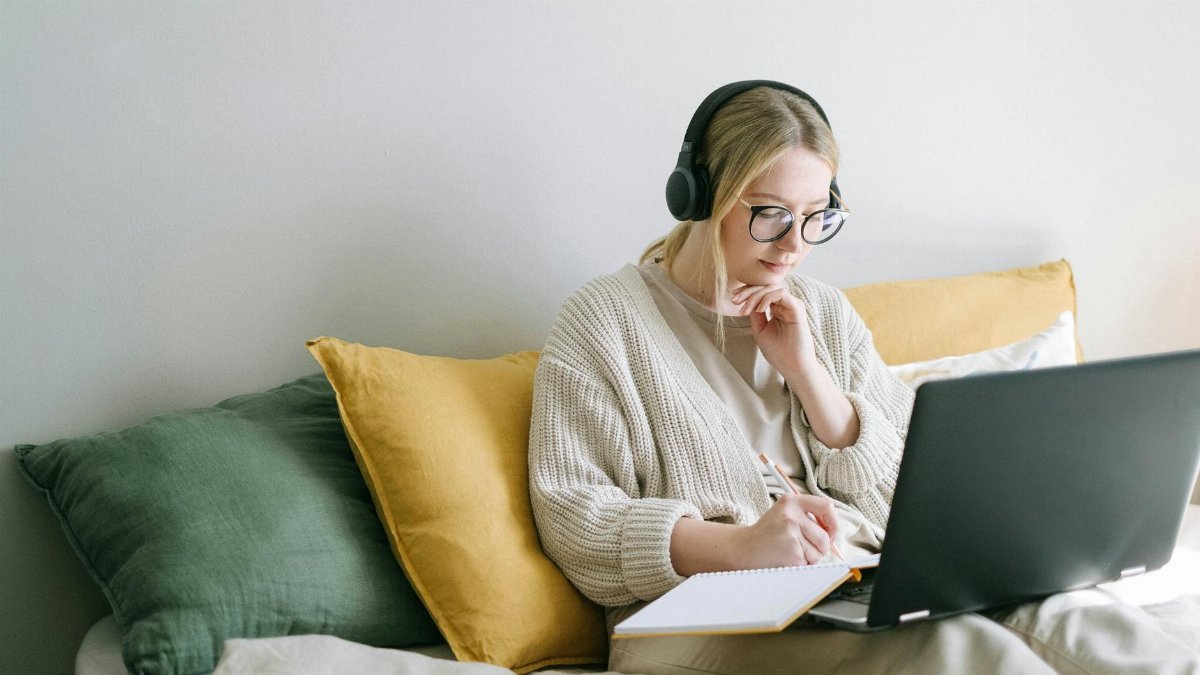 A focused woman in glasses and headphones works on a laptop from a cozy bed.