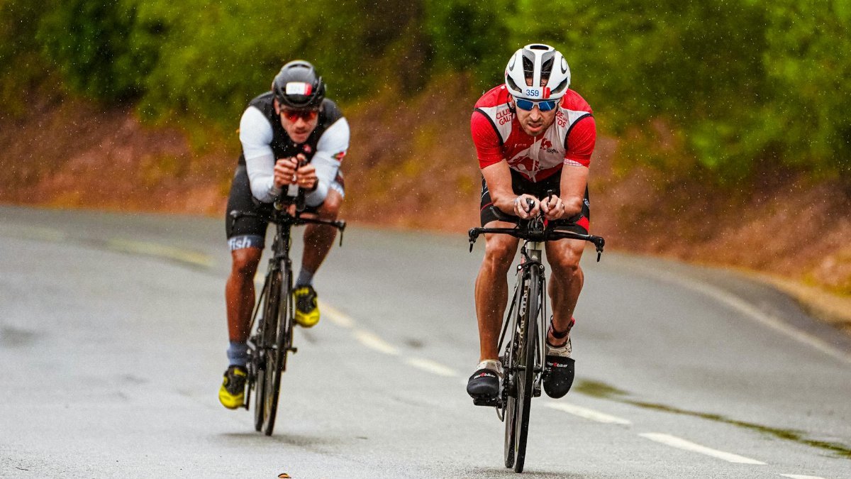 Full body of concentrated young tanned sportsmen riding bicycles on uneven asphalt road under rain in daytime