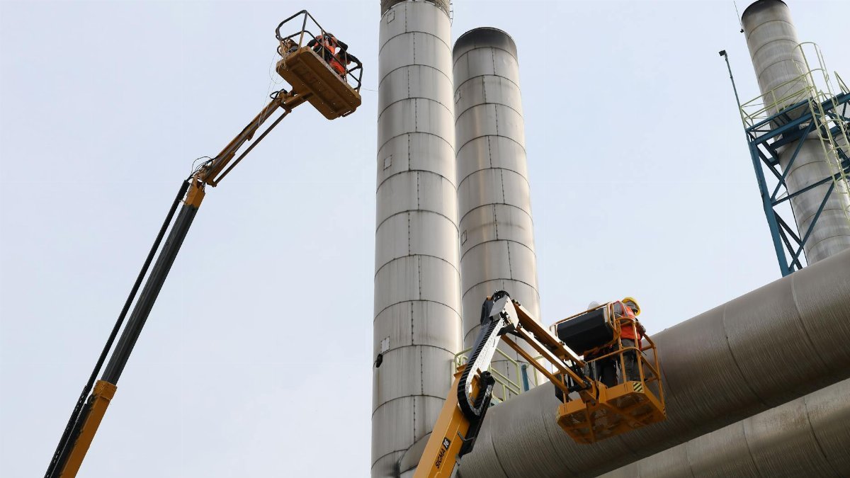 Workers using lifts for maintenance near towering factory chimneys against a clear sky.