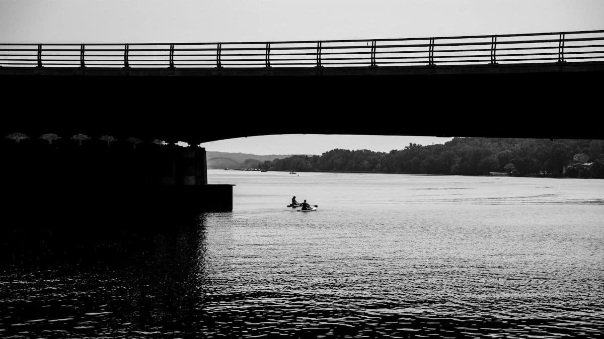 Black and white image of kayakers paddling under an urban bridge, evoking leisure and exploration.