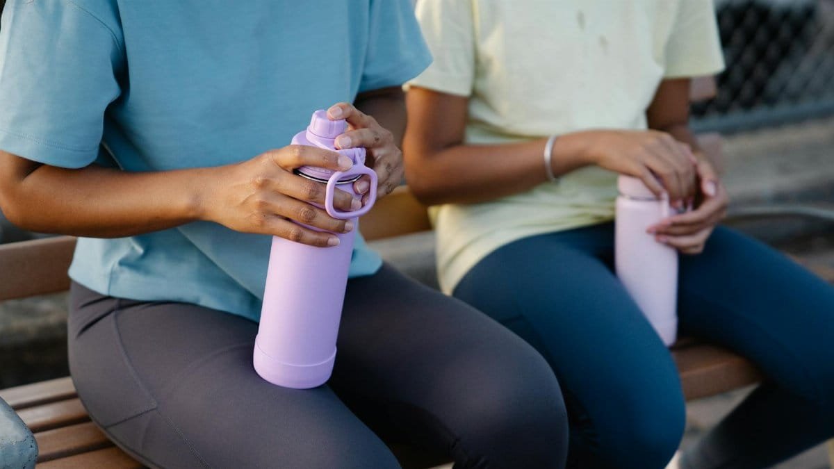 Two women in casual outfits seated outdoors, holding water bottles, taking a break on a wooden bench.