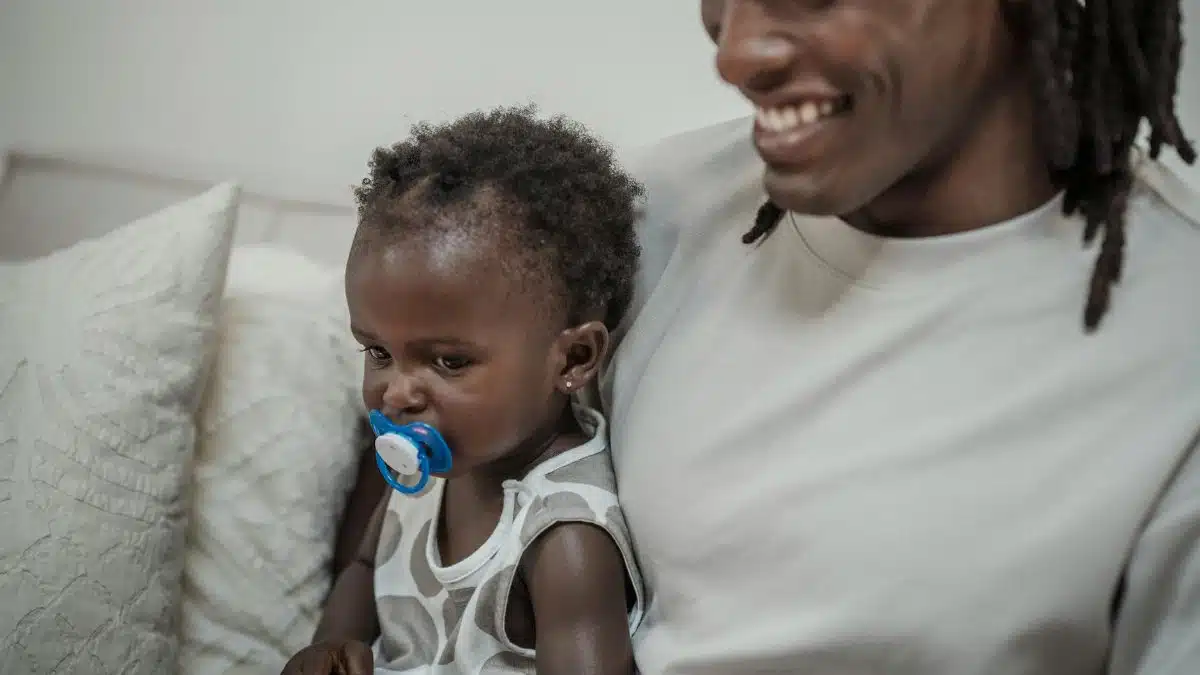 A smiling father holding his daughter with a soother, sitting together indoors.