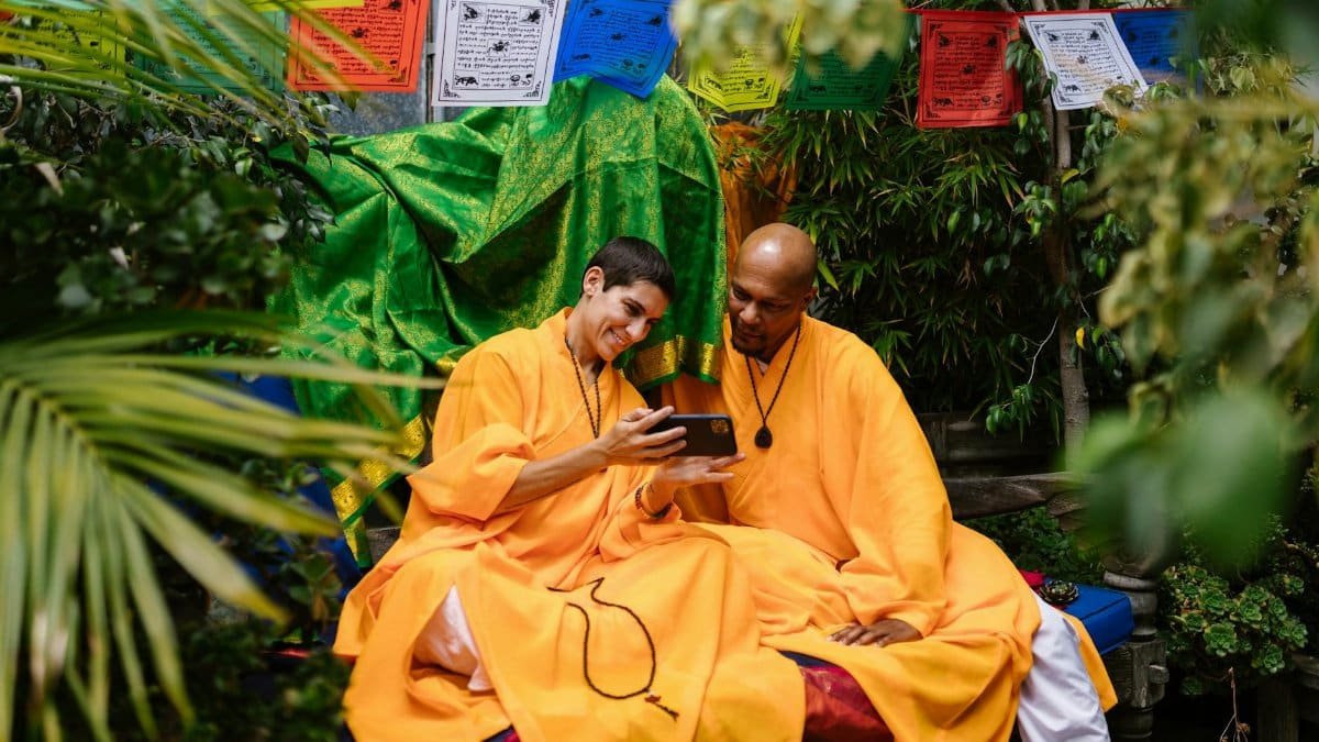 Two monks in bright robes share a moment with smartphone, surrounded by lush greenery and colorful prayer flags.