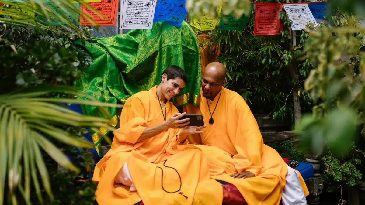 Two monks in bright robes share a moment with smartphone, surrounded by lush greenery and colorful prayer flags.