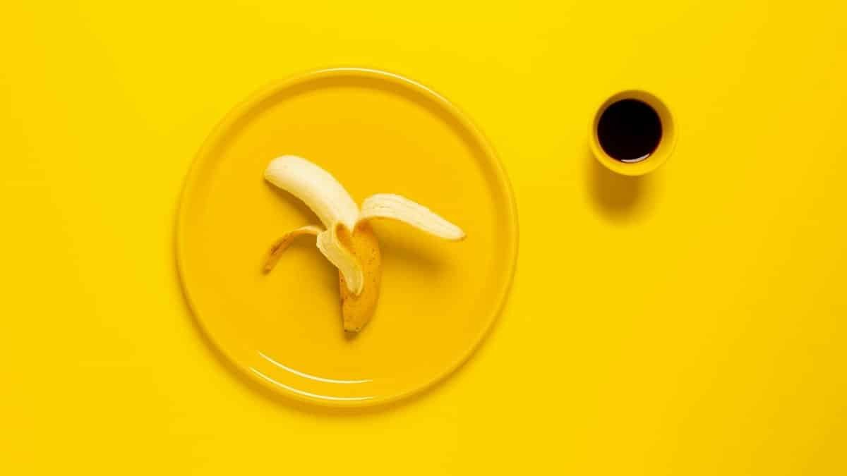 A peeled banana on a yellow plate beside a cup of black coffee over a vibrant yellow background.