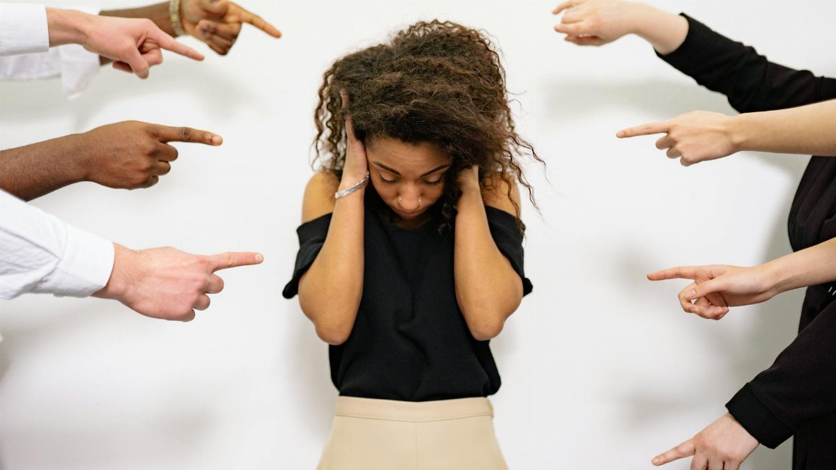 A young woman with curly hair looks distressed as multiple hands point accusatory fingers at her.