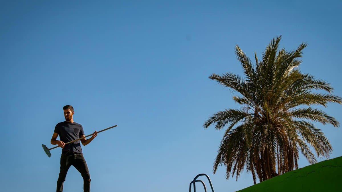 A man stands on a hill with a broom, under a clear blue sky next to a palm tree.