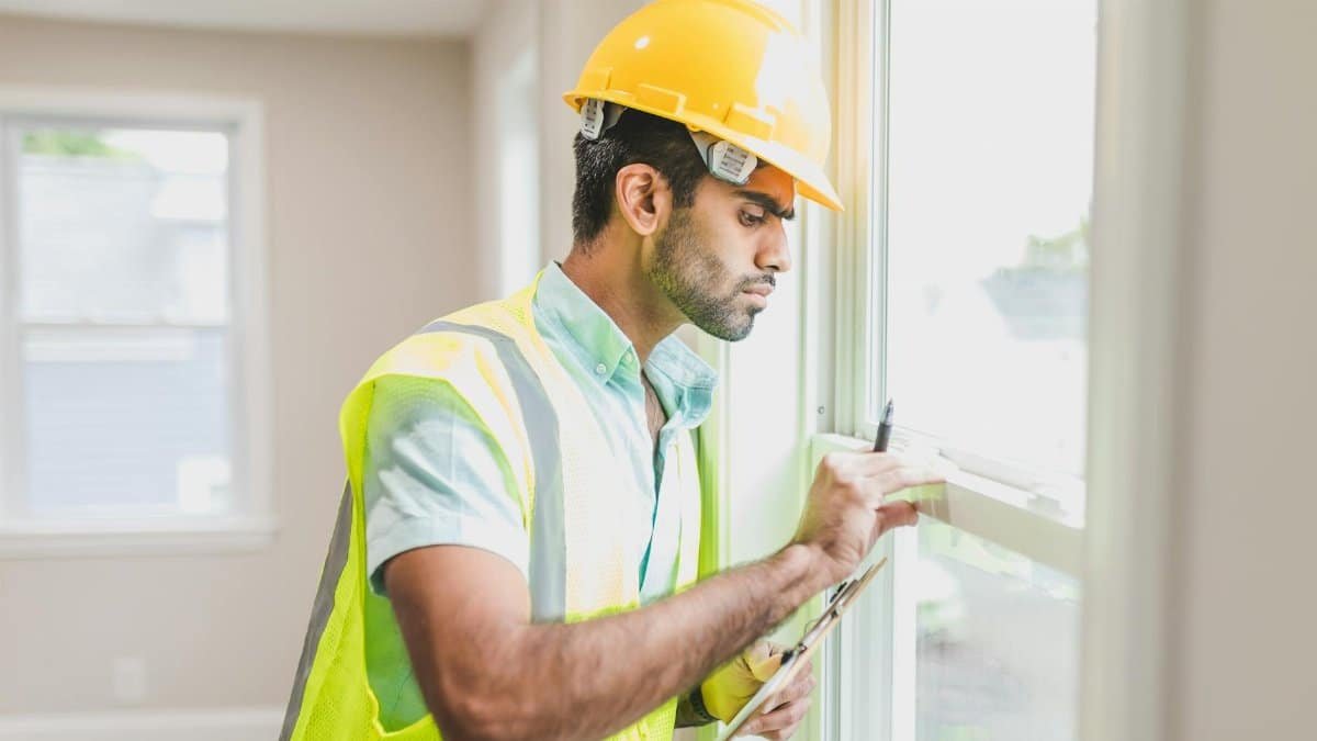 Construction worker with hard hat and vest inspecting window indoors, side view.