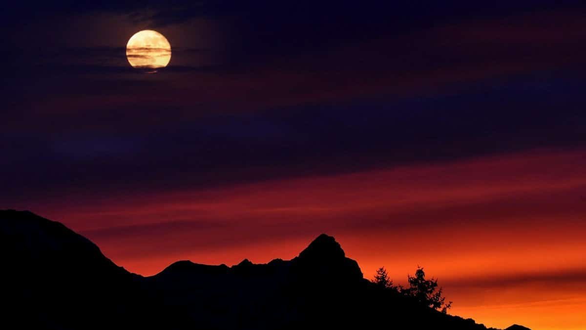 A captivating scene of a full moon above silhouetted mountain peaks against a vivid sunset sky.