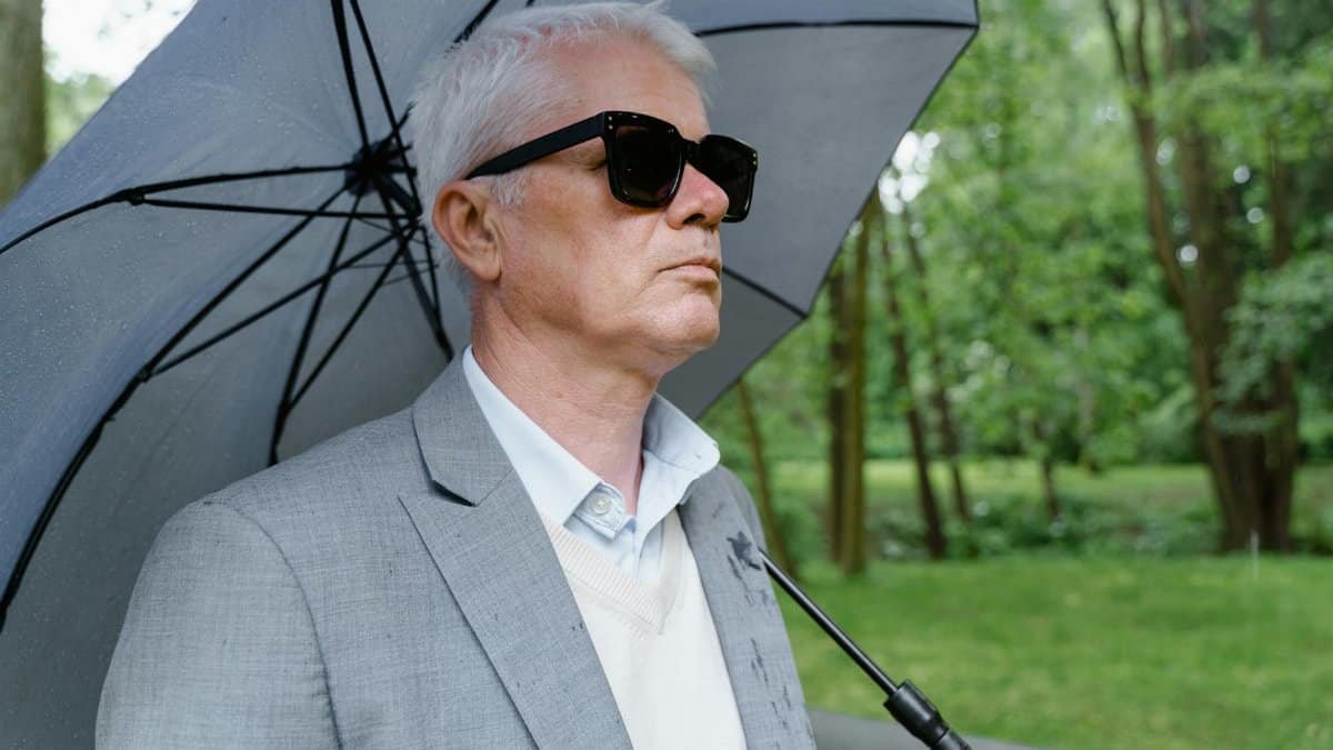Senior man with white hair wears sunglasses and holds an umbrella in a park during rain.