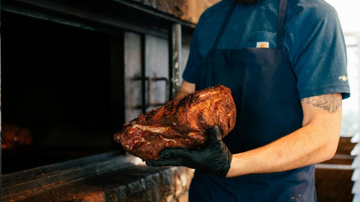 Chef holding seasoned meat with gloves near barbecue pit, cooking outdoors.