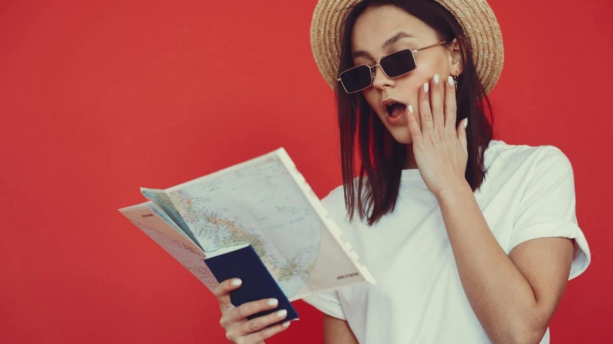A trendy woman in sunglasses and a hat reads a map with surprise against a red background.