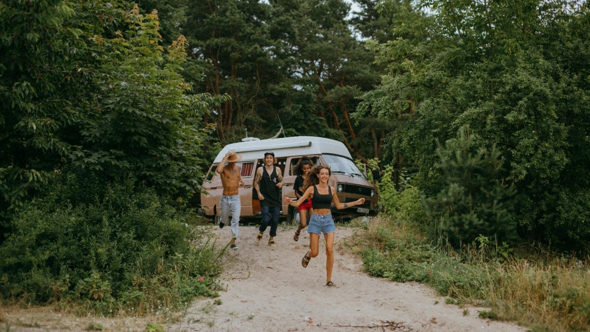 Group of friends running joyfully from a camper van in a lush forest setting during summer.