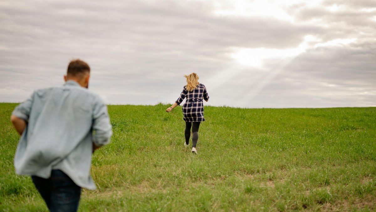 A carefree couple runs joyfully through a green meadow under an overcast sky, embodying freedom and adventure.