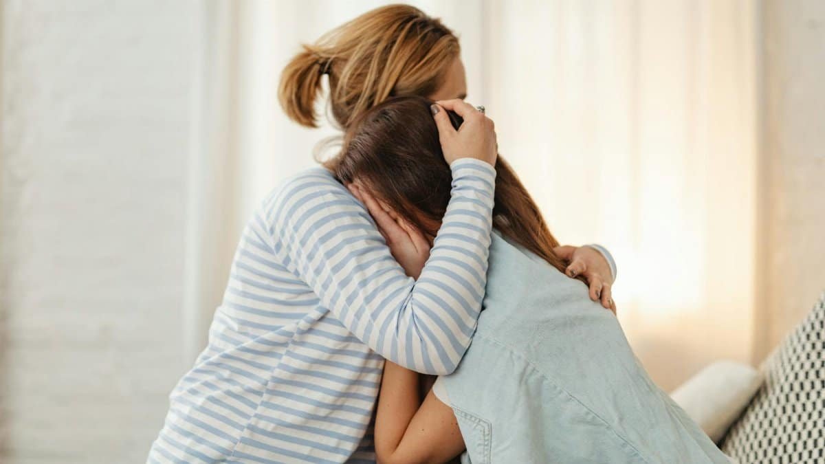 A mother lovingly comforts her teenage daughter in an emotional embrace indoors.