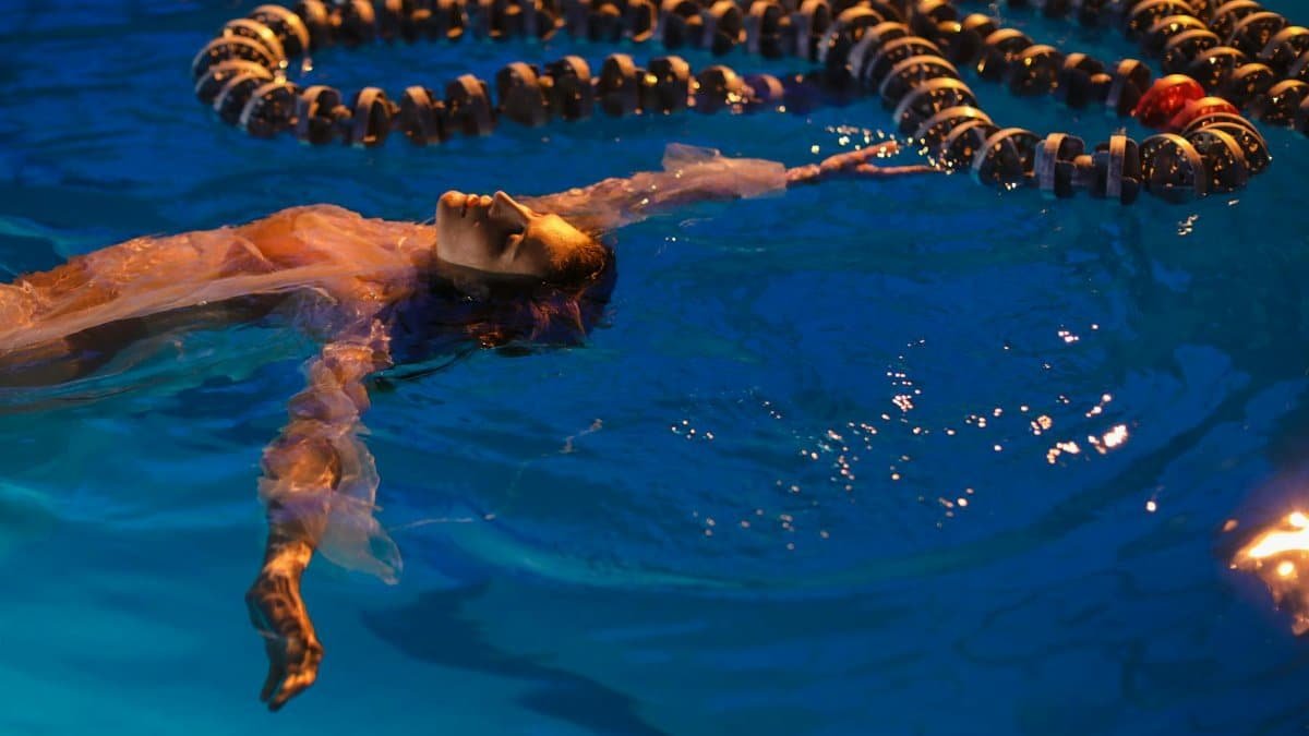 A woman floats calmly in a swimming pool under evening lights, surrounded by lane dividers.