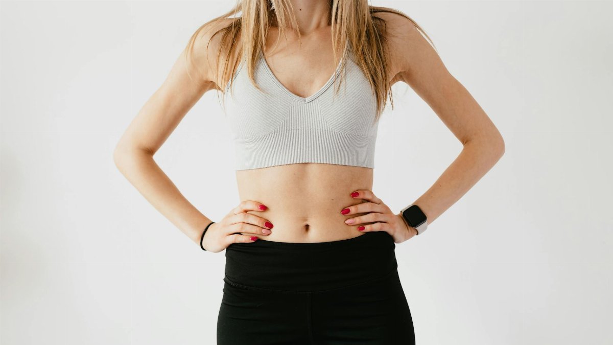 Faceless slim anonymous blond female in sports bra and black leggings in wearable bracelet showing perfect belly on white background while standing with hands on waist