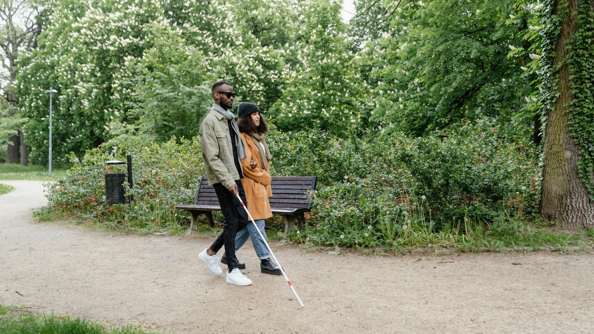 A visually impaired couple walks hand-in-hand with a cane through a verdant park path.