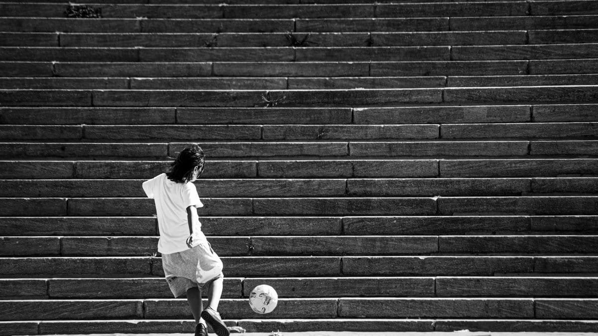 Black and white photo of a child kicking a soccer ball on stone steps, urban setting.