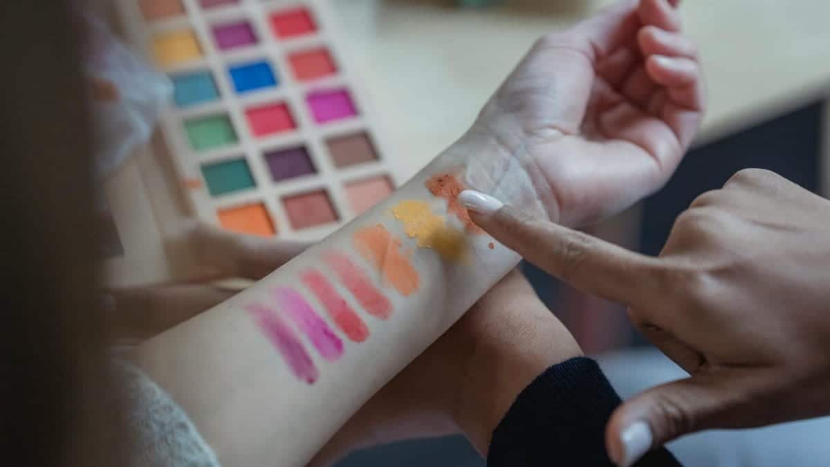 From above of crop unrecognizable young female makeup artist pointing at arm of client while applying colorful eyeshadows on wrist for testing