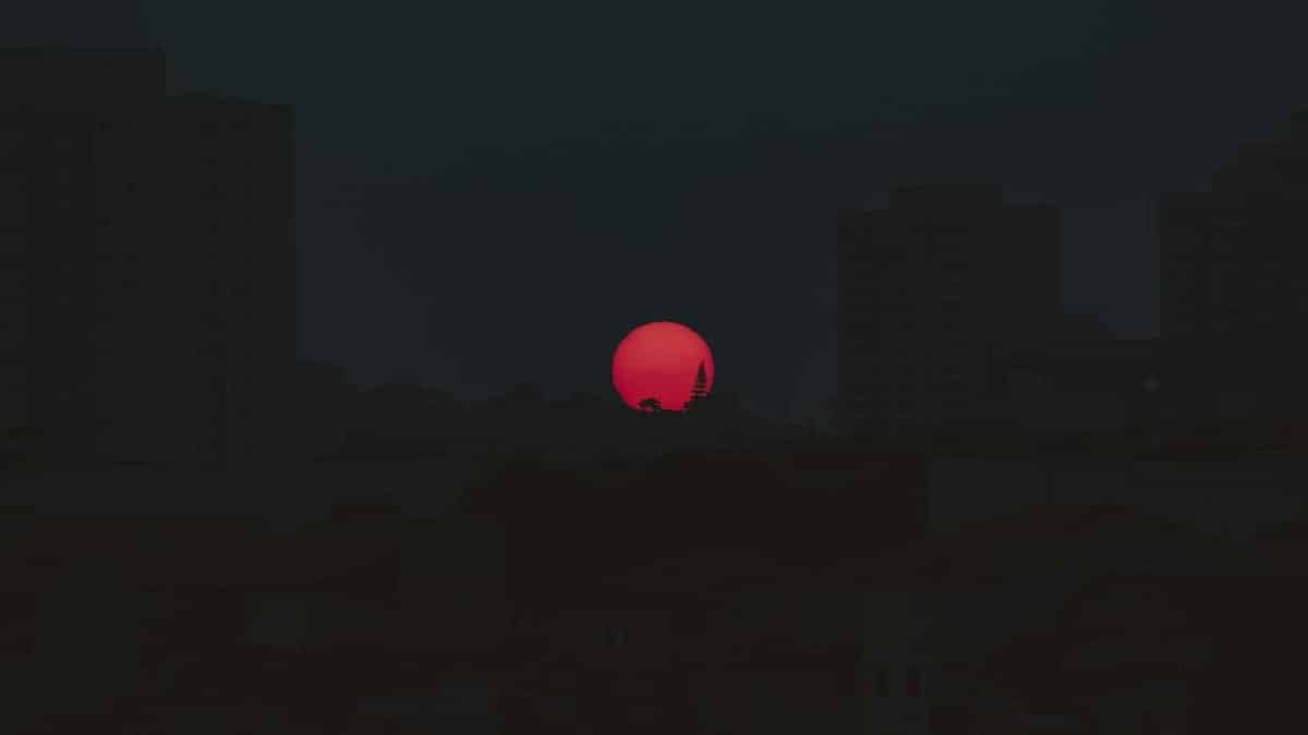 A vibrant red sun sets behind a silhouetted cityscape creating a dramatic evening scene.