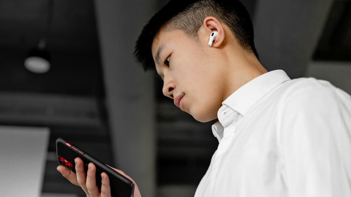Side view of a young man wearing wireless earbuds and using a smartphone indoors.
