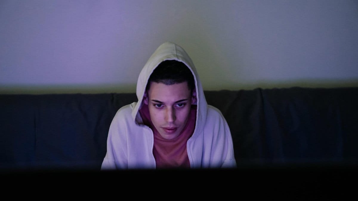 Focused young ethnic male in hood looking forward while sitting on couch in house room