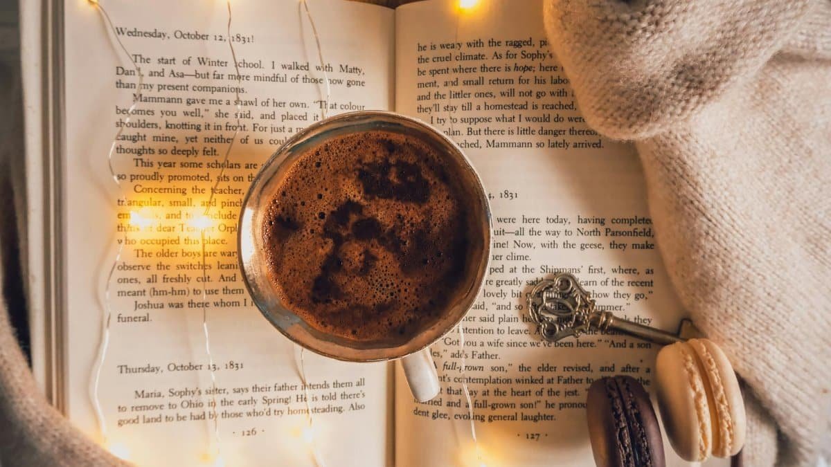 Warm coffee cup on an open book with macarons and fairy lights for a cozy reading ambiance.